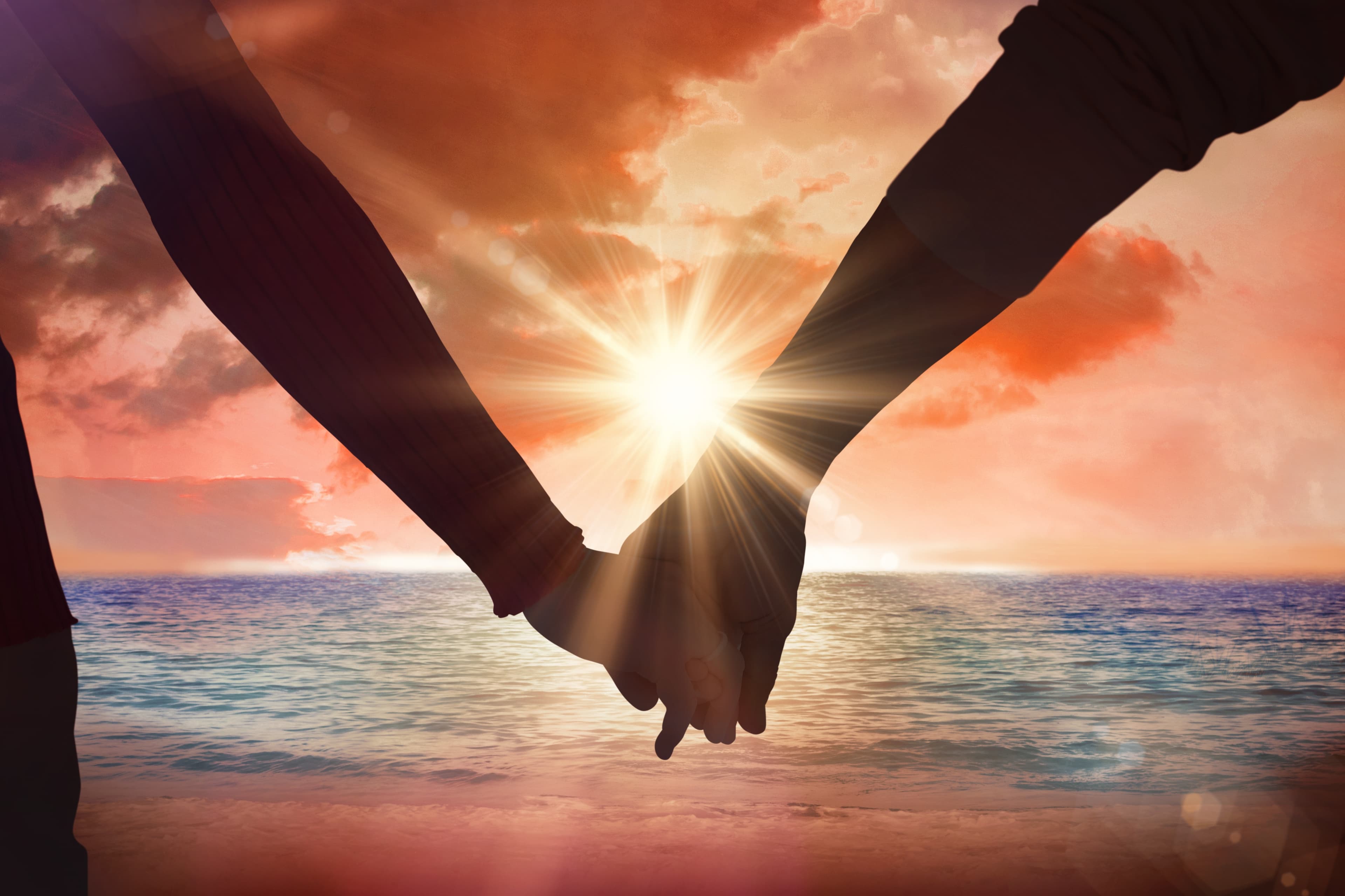 Silhouetted couple holding hands against a bright sunburst and orange sunset over the ocean.
